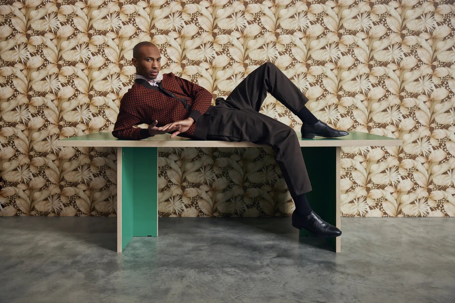 A person in stylish clothing reclines on a HKLIVING Dining Table, Rectangular, 220cm by HKLIVING, set against floral-patterned wallpaper with one leg raised expressively. The table sits on a gray concrete floor.