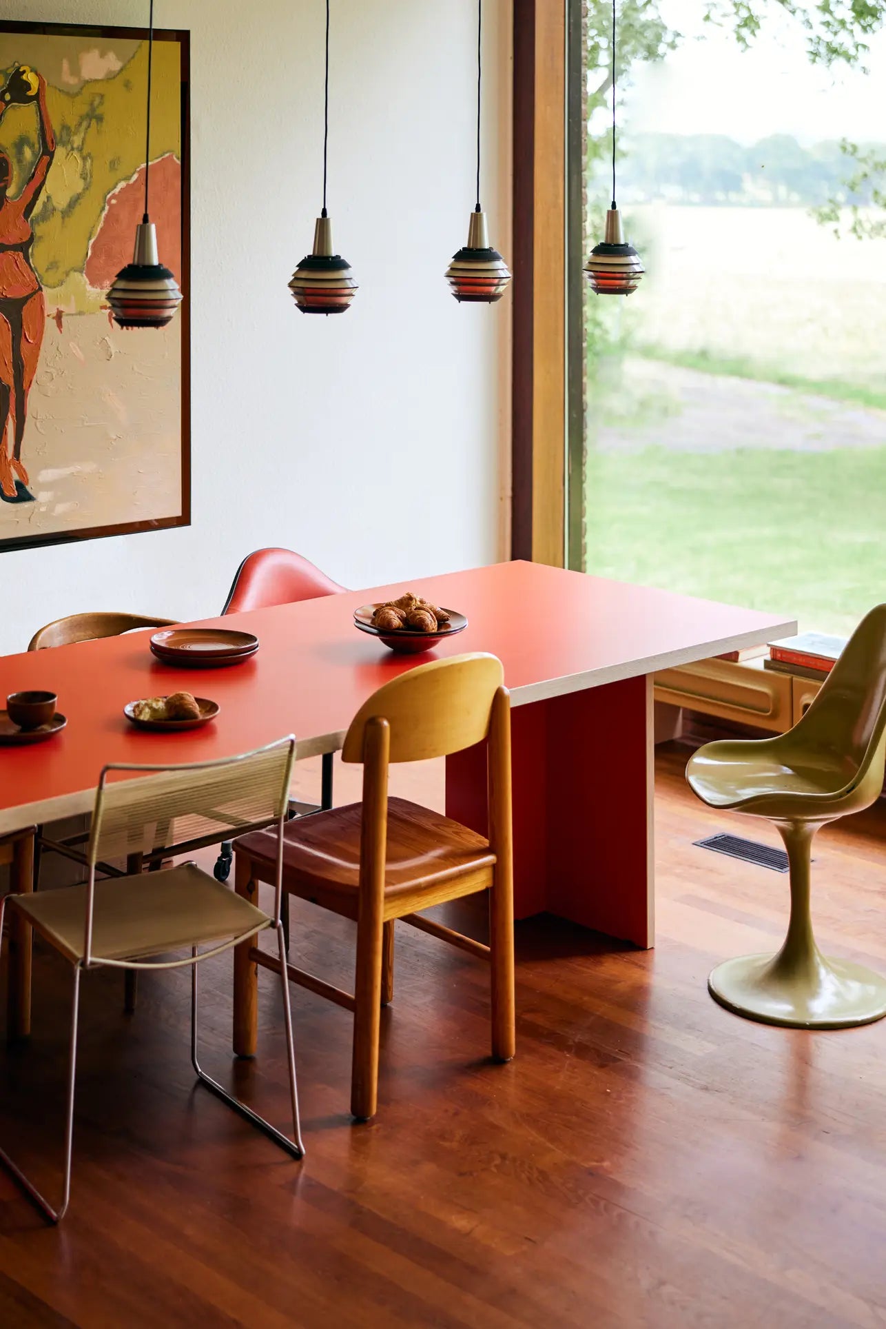 A modern dining room features the HKLIVING Dining Table, Rectangular, 220cm by HKLIVING with assorted chairs and pendant lights. Art decorates the white wall, large windows offer a grassy view, and plates and cups are set on the table.