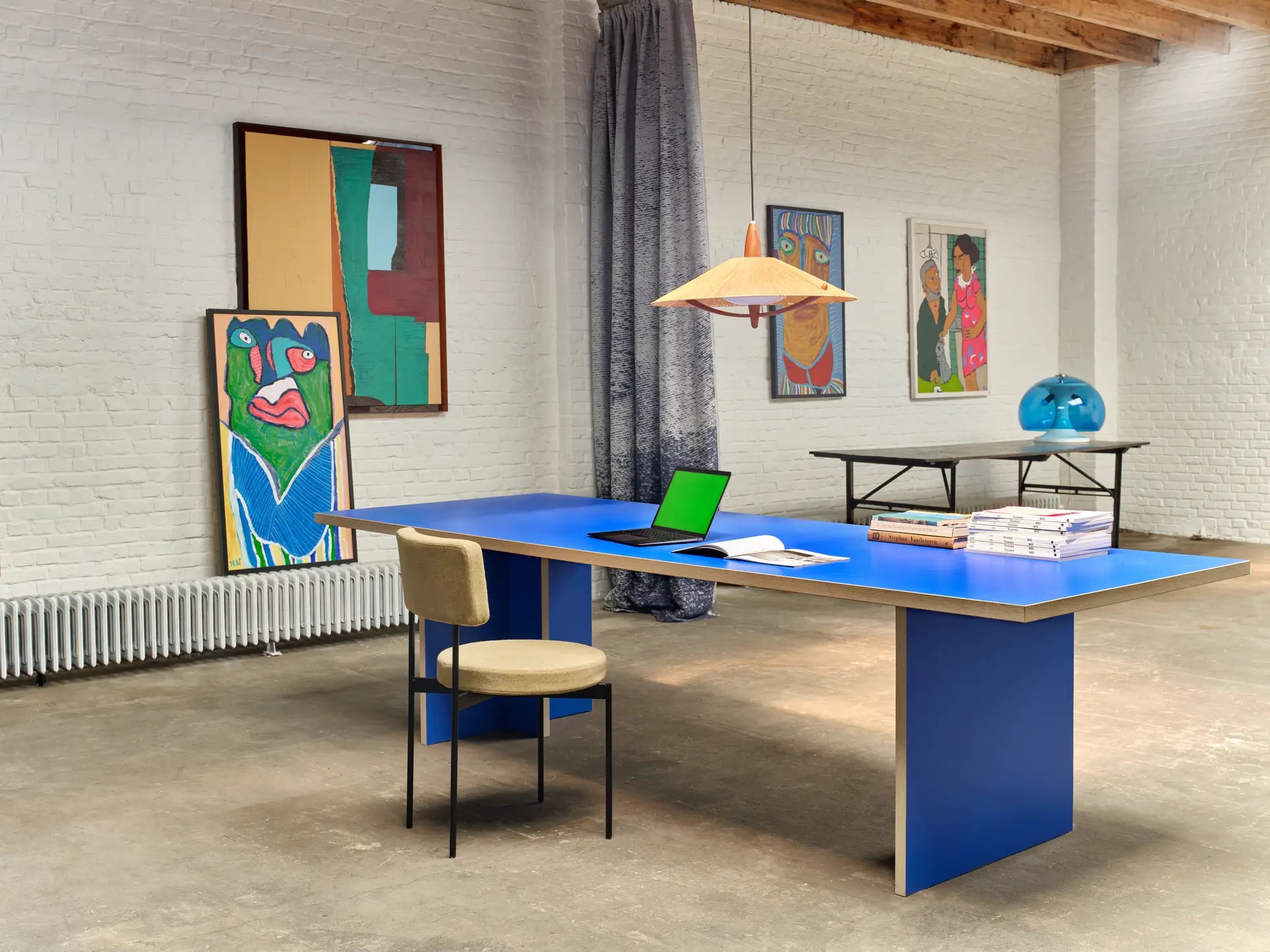 A modern office features a bright blue desk, beige chair, laptop, books, and bold artwork on white brick walls. A pendant lamp hangs above the HKLIVING Dining Table, Rectangular, 280cm by HKLIVING under exposed ceiling beams.