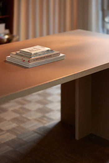A stack of books rests on the HKLIVING Dining Table, Rectangular, 280cm by HKLIVING in a warmly lit room with parquet flooring and soft curtains, reflecting timeless design.