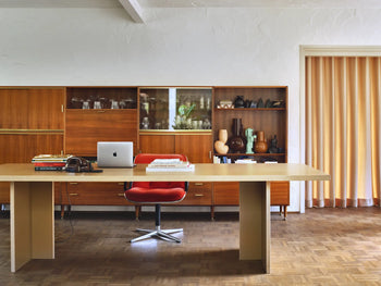A modern office with a tan HKLIVING Dining Table, Rectangular, 280cm as a desk, red swivel chair, laptop, books, and headphones. Wooden cabinets with glassware and vases stand behind; parquet flooring and striped curtains complete the timeless look.