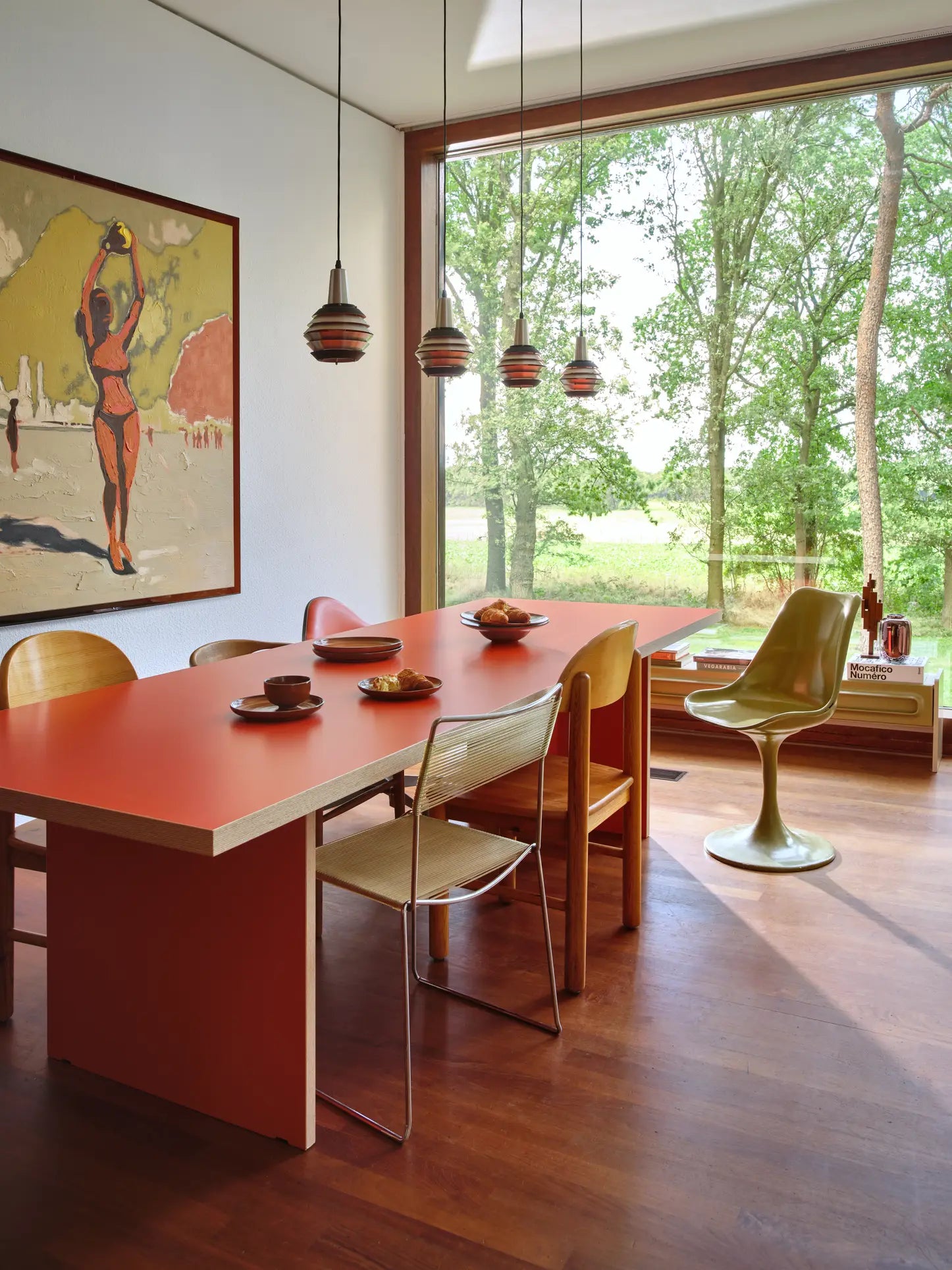 A mid-century modern dining room features a timeless design with the HKLIVING Dining Table, Rectangular, 280cm in red, four wooden chairs, a swimwear portrait painting, retro pendant lights, tree views through a large window, and a yellow swivel chair by a low table.