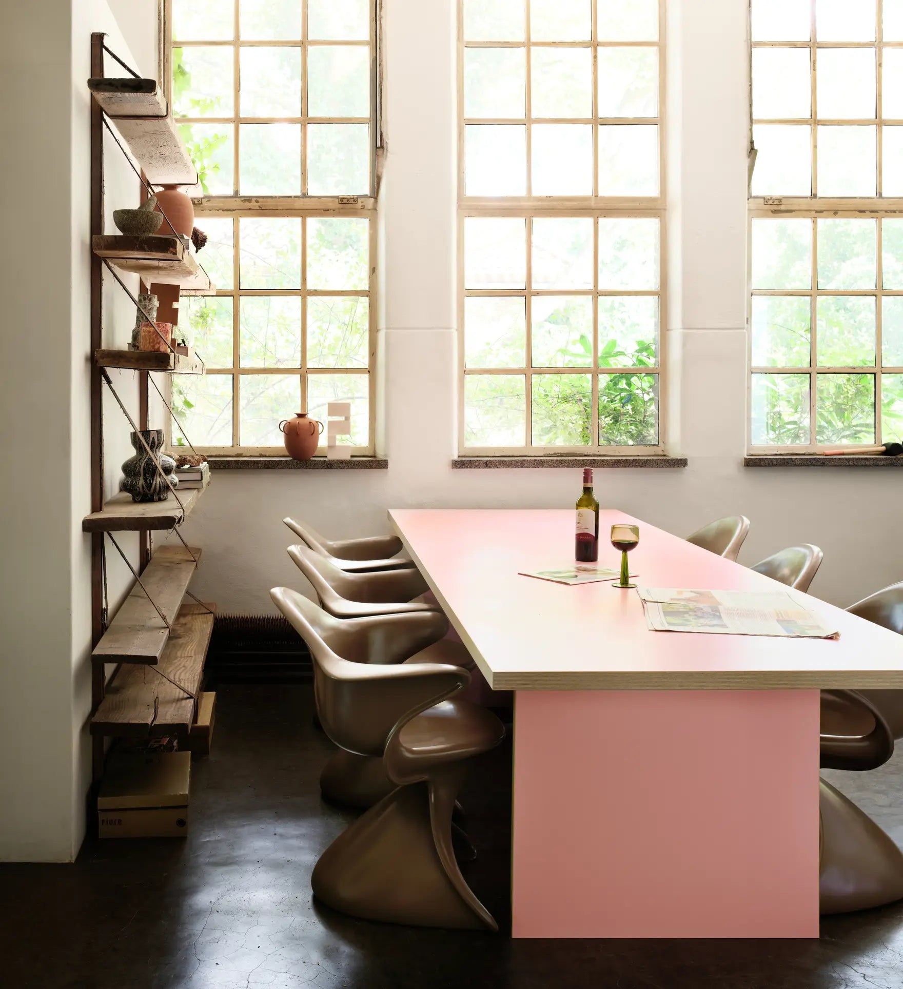 A modern dining area features the HKLIVING Dining Table, Rectangular, 280cm with a pink base, surrounded by curvy beige chairs. Large windows brighten the space, and a tall wooden shelf displays plants and decor. A wine bottle and glass sit on the table.
