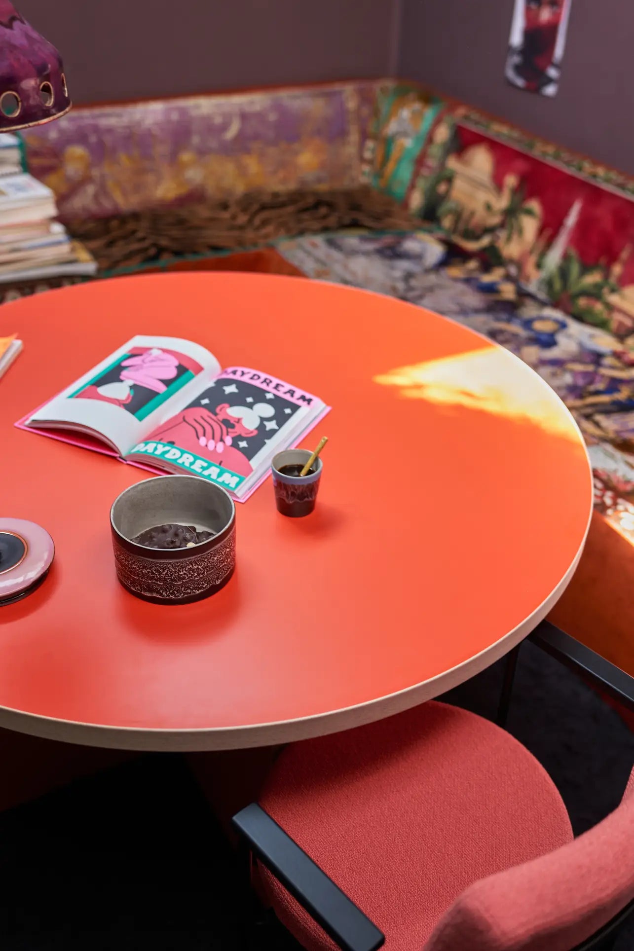 The HKLIVING Dining Table, Round, 130cm in vibrant orange displays a magazine, bowl, cup, and pen. Behind it, a cushioned bench with colorful patterned upholstery and stacked books adds charm to the space.