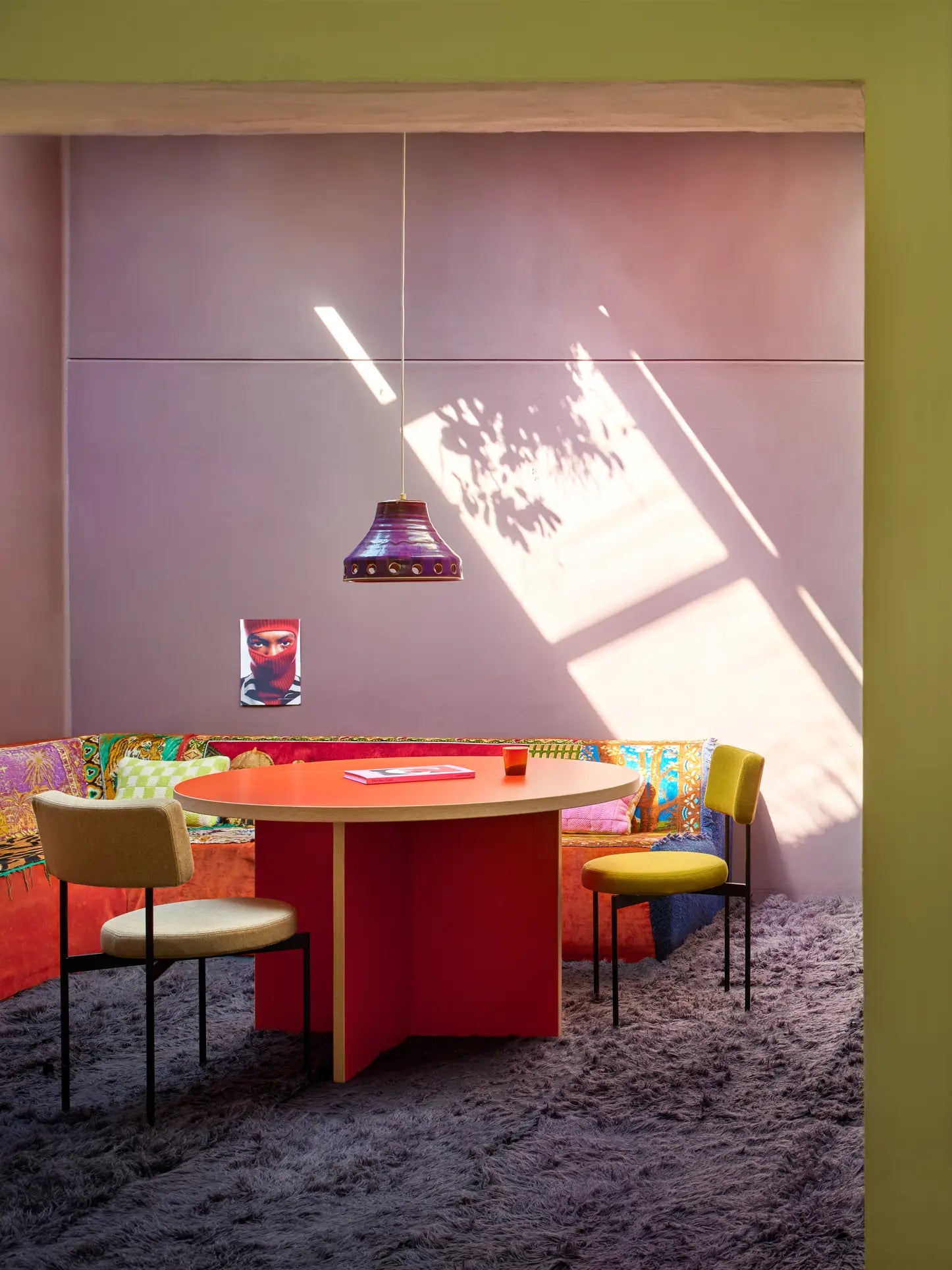A sunlit room features a round HKLIVING Dining Table (130cm), three chairs, a colorful corner sofa, patterned rug, purple hanging lamp, and a poster on a violet wall. Sunlight streams in through the large window, casting soft shadows.