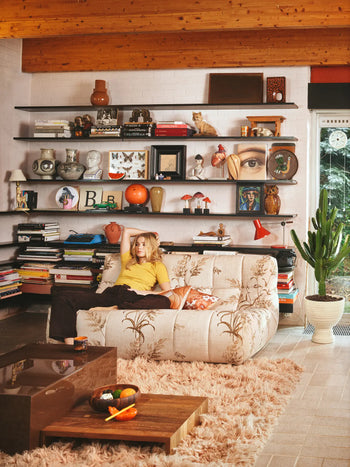 A woman relaxes on the HKLIVING Lazy Lounge Bench in a cozy living room filled with books, art, and decor. A cactus, fruit-topped coffee table, fluffy rug, and wooden ceiling beams complete the eclectic, warm vibe.