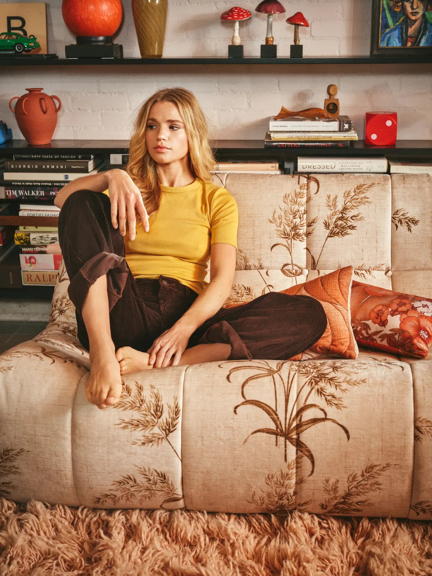 A woman with long blonde hair, in a yellow top and dark pants, sits barefoot on the HKLIVING Lazy Lounge Bench in a stylish room with shelves, books, red pottery, and decorative decor by HKLIVING visible in the background.