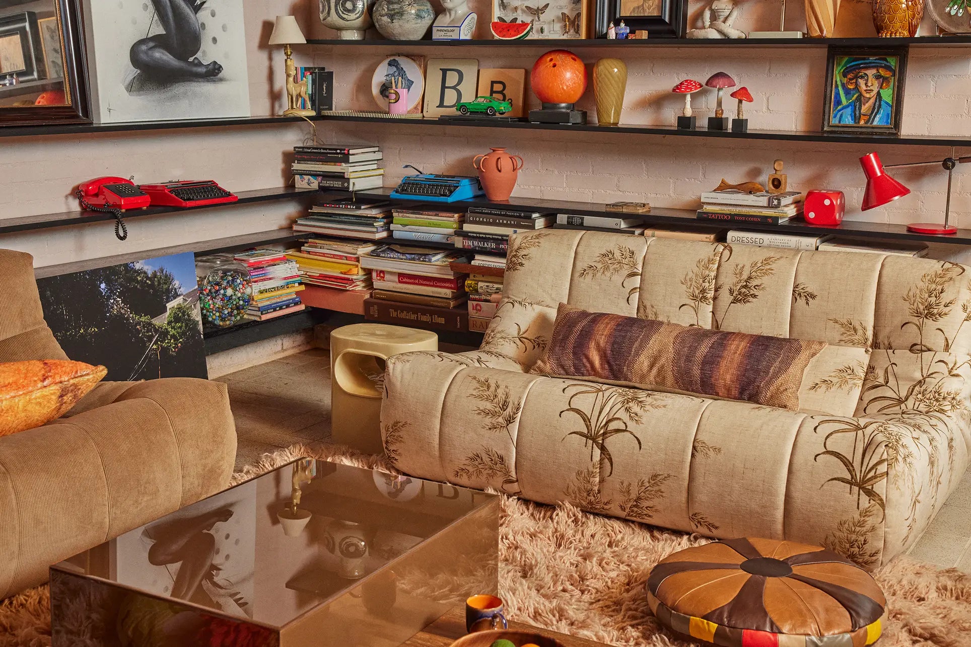 A cozy living room features a patterned beige sofa, the HKLIVING Lazy Lounge Chair Corduroy by HKLIVING, a furry rug, and a glass coffee table. Wall shelves display books, art, vintage items, and colorful decor for an eclectic retro style.