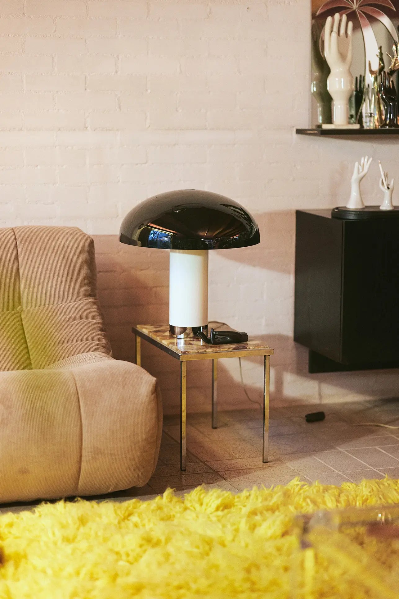 The HKLIVING Lazy Lounge Chair Corduroy in beige sits by a small glass side table with a black and white mushroom lamp, atop a yellow shag rug. Hand sculpture decor accents a shelf set against a white brick wall.