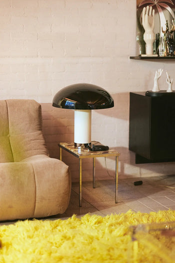 The HKLIVING Lazy Lounge Chair Corduroy in beige sits by a small glass side table with a black and white mushroom lamp, atop a yellow shag rug. Hand sculpture decor accents a shelf set against a white brick wall.