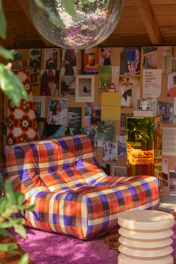 A cozy room features the HKLIVING Lazy Lounge Chair, playful art and fashion magazine prints on the wall, a white round side table, a purple rug, and a disco ball hanging from the ceiling.