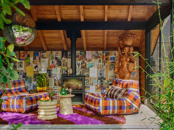 A colorful, retro living room features playful prints, an HKLIVING Lazy Lounge Chair, round side table with fruit, wood stove, shaggy purple rug, disco ball, wooden ceiling, eclectic art and photos on the wall, and a wooden statue.