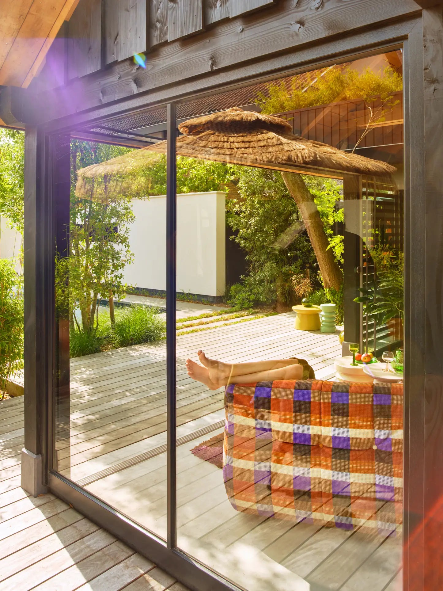 A person relaxes indoors with feet on the HKLIVING Lazy Lounge Chair, wrapped in a colorful blanket. Sunlight streams through the glass door, reflecting an outdoor wooden deck and lush greenery for a peaceful HKLIVING ambiance.