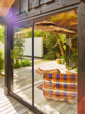 A person relaxes indoors with feet on the HKLIVING Lazy Lounge Chair, wrapped in a colorful blanket. Sunlight streams through the glass door, reflecting an outdoor wooden deck and lush greenery for a peaceful HKLIVING ambiance.