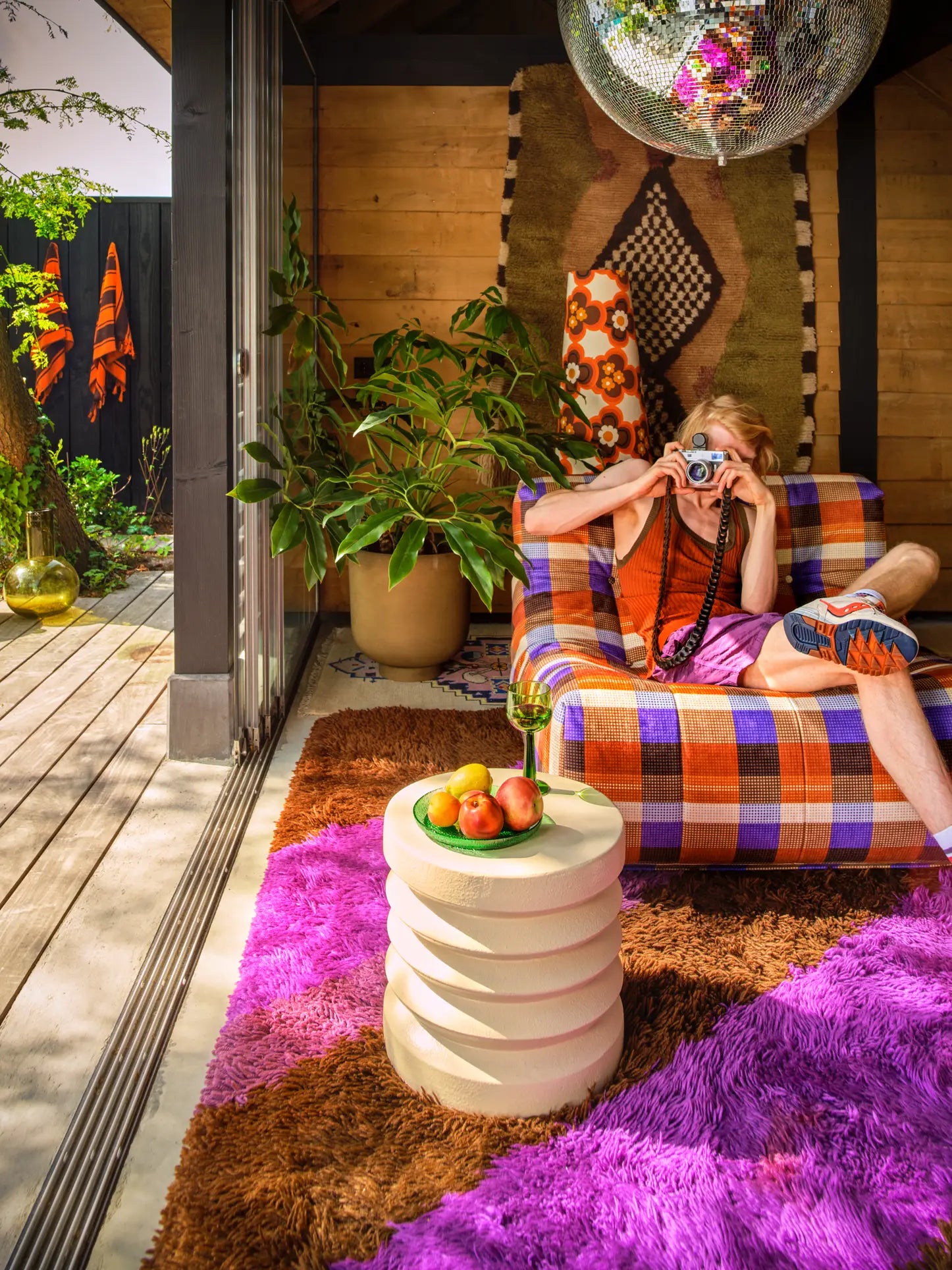 A person in colorful clothing sits indoors on the HKLIVING Lazy Lounge Chair by HKLIVING, surrounded by playful prints, a purple shag rug, mirrored disco ball, plants, and a small table with fruit and a drink.