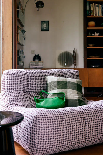 A modern living room featuring the HKLIVING Lazy Lounge Chair in houndstooth, topped with a green checkered pillow and a bright green bag. Playful prints and wooden shelves with decor add stylish touches to the background.