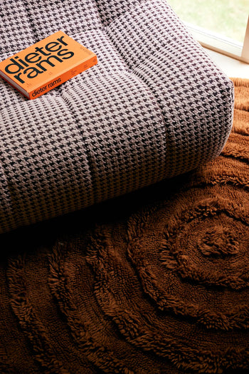 An orange Dieter Rams book sits on an HKLIVING Lazy Lounge Chair with playful prints, next to a brown rug with circular patterns, by a sunlit window.