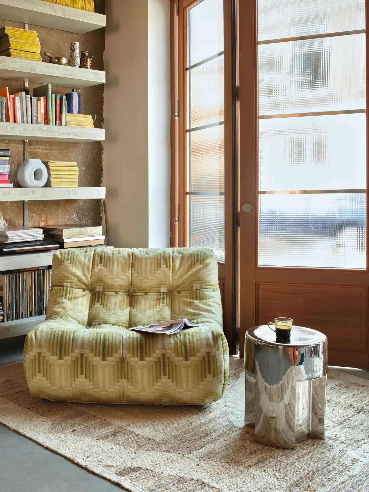 The HKLIVING Lazy Lounge Chair with a green playful pattern sits on a textured rug next to a metallic side table with a glass, while bookshelves and large frosted windows brighten the cozy space with natural light.