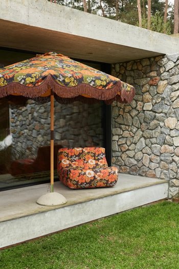 The HKLIVING Lazy Lounge Chair Outdoor, featuring a floral pattern and matching umbrella, is placed on a concrete patio next to a stone wall, overlooking a grassy lawn and surrounded by trees outside a modern building.