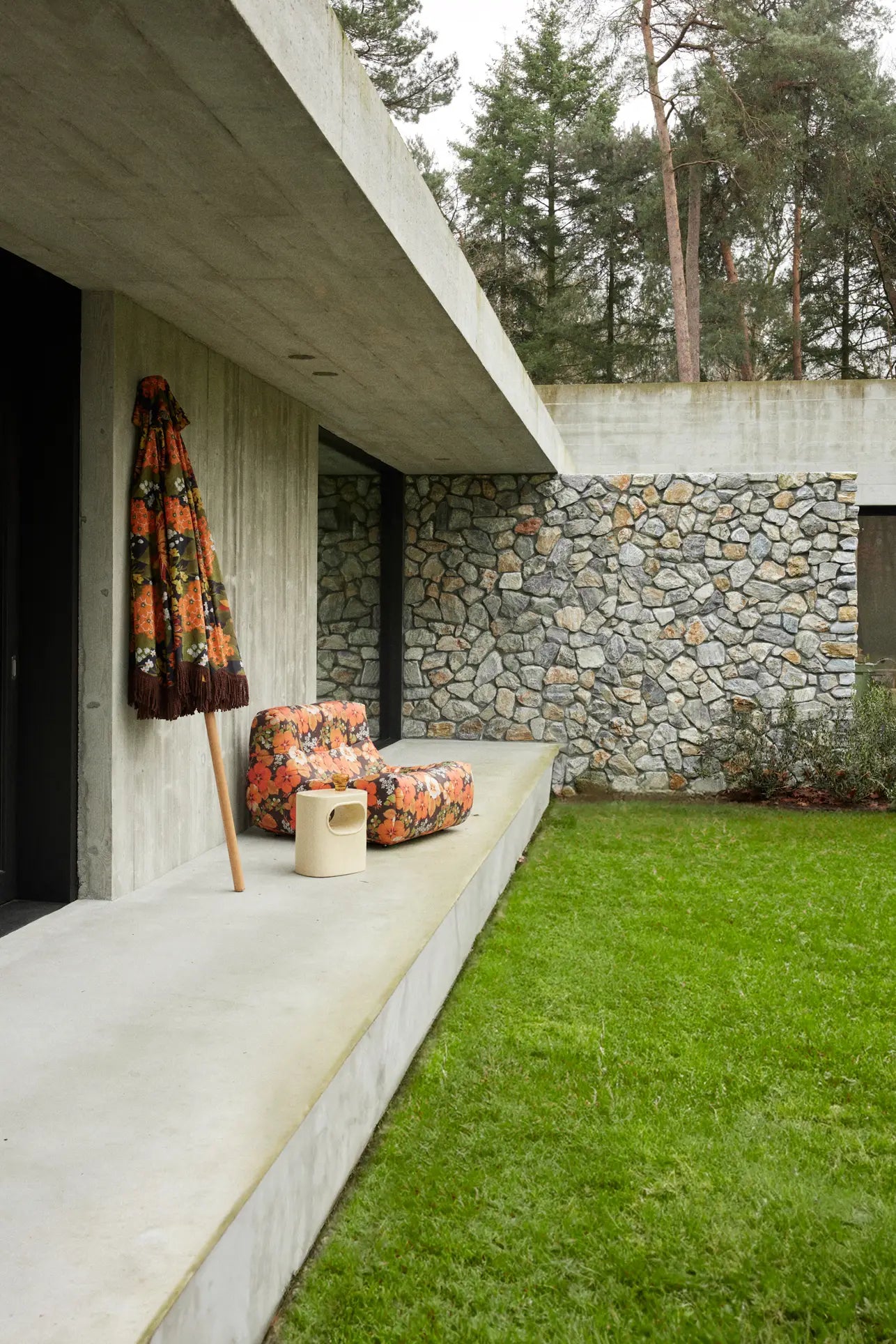 A modern concrete house features a stone wall, wide patio, grassy lawn, and the HKLIVING Lazy Lounge Chair Outdoor by HKLIVING with a floral umbrella. Tall trees create a scenic backdrop.
