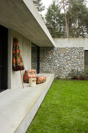 A modern concrete house features a stone wall, wide patio, grassy lawn, and the HKLIVING Lazy Lounge Chair Outdoor by HKLIVING with a floral umbrella. Tall trees create a scenic backdrop.