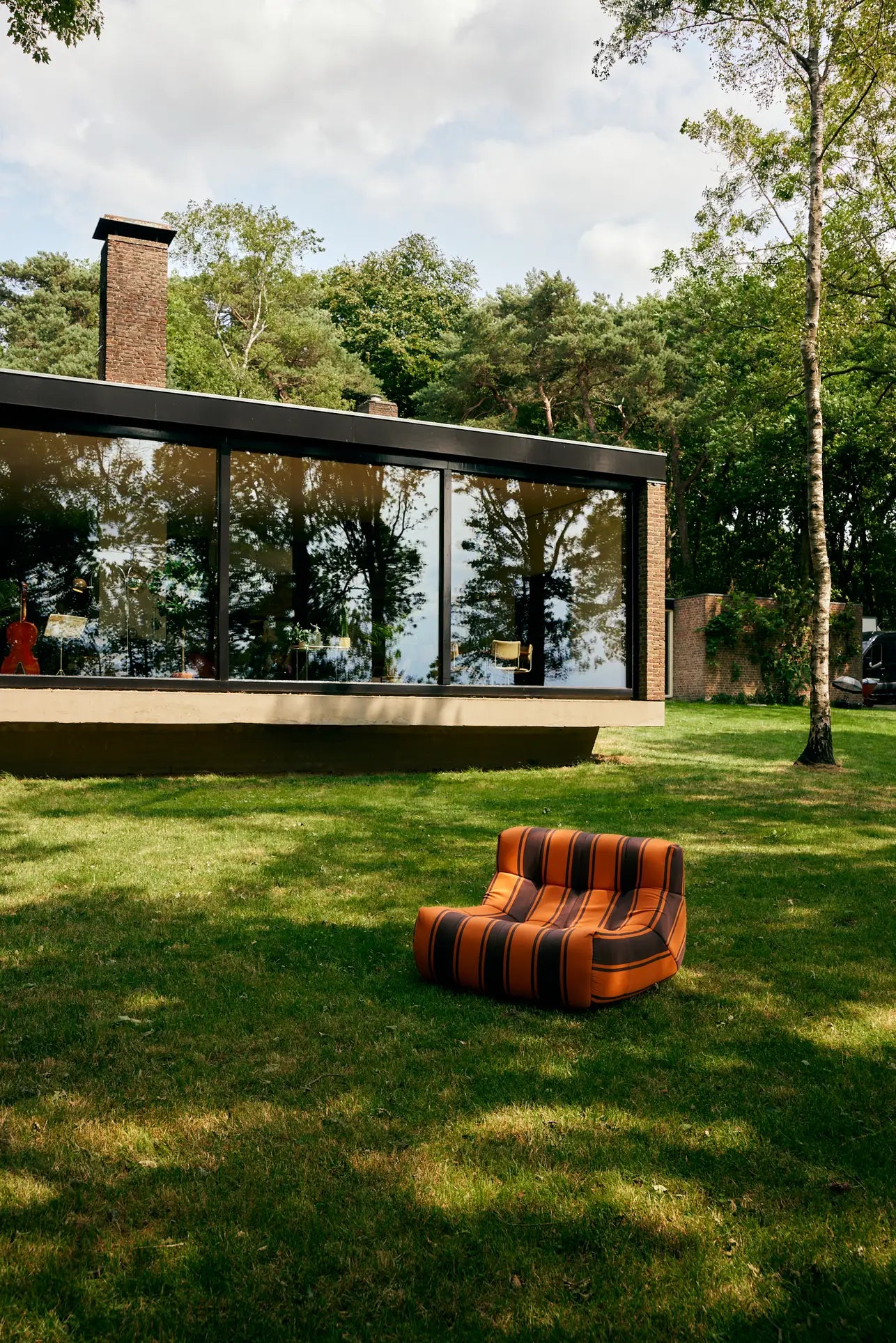 The HKLIVING Lazy Lounge Chair Outdoor by HKLIVING, with retro orange and brown stripes, sits on a grassy lawn outside a modern glass-walled house, surrounded by trees under a partly cloudy sky.