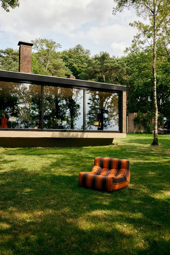 The HKLIVING Lazy Lounge Chair Outdoor by HKLIVING, with retro orange and brown stripes, sits on a grassy lawn outside a modern glass-walled house, surrounded by trees under a partly cloudy sky.