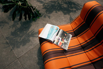 An open magazine rests on the orange and brown striped cushion of the HKLIVING Lazy Lounge Chair Outdoor by HKLIVING, set on a gray tiled floor with plant shadows and leafy details in the background.
