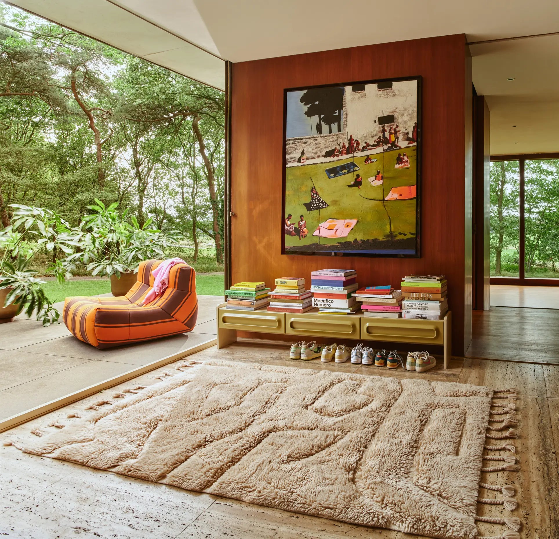 Modern living space with large windows, green plants, the HKLIVING Lazy Lounge Chair Outdoor, a textured beige rug, low shelves of colorful books, neatly lined shoes, wall art of people relaxing on grass, and lush trees outside.