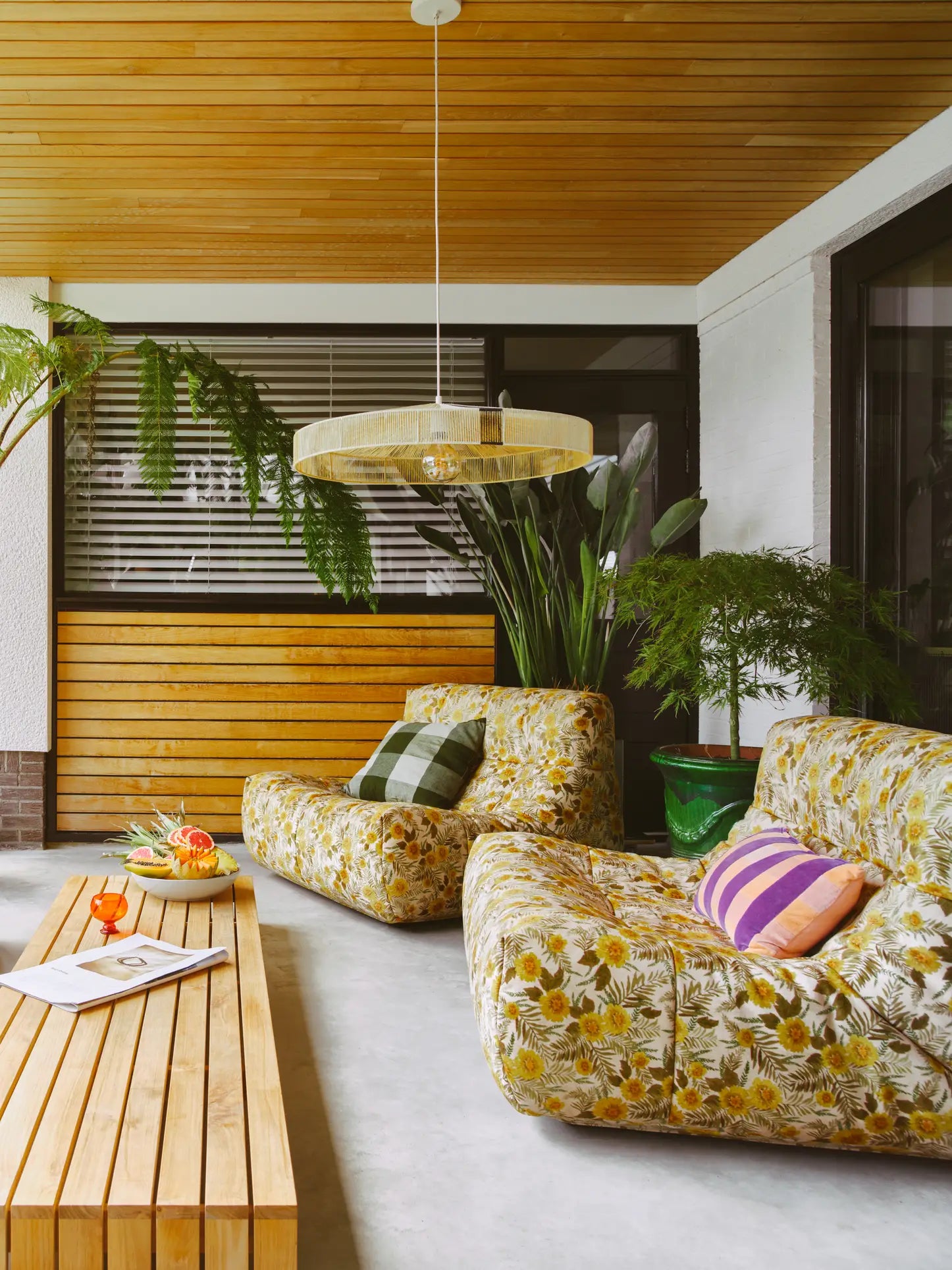 A cozy patio with HKLIVING Lazy Lounge Chairs featuring playful yellow floral prints, a wooden table topped with drinks, fruit, and a magazine, potted plants, and a modern pendant light. Wood paneling and windows complete the inviting scene.