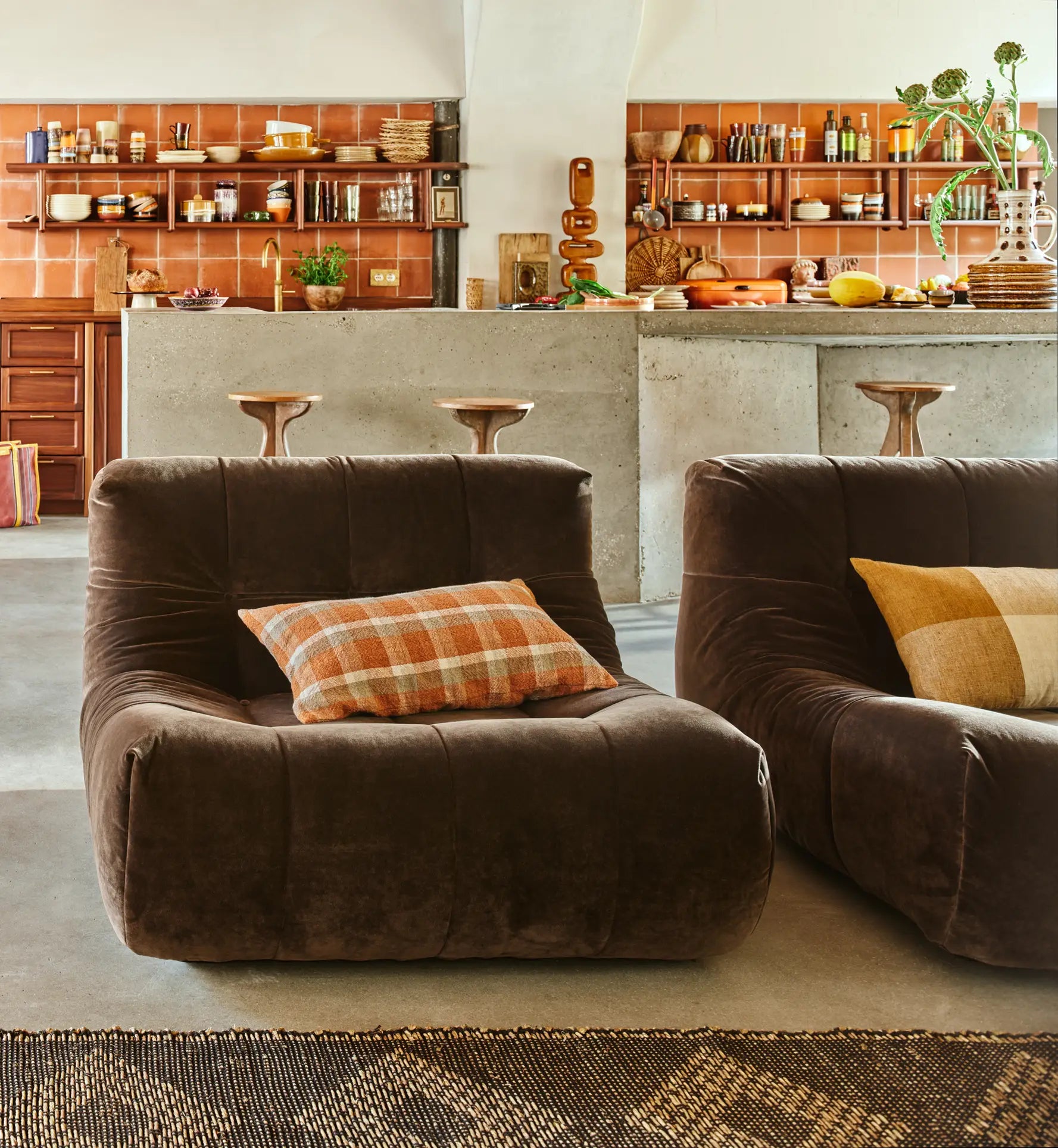 A cozy kitchen featuring HKLIVING Lazy Lounge Chairs with plaid pillows in the foreground, and a concrete island with wooden stools and open shelves displaying dishes and cookware in the background.