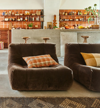A cozy kitchen featuring HKLIVING Lazy Lounge Chairs with plaid pillows in the foreground, and a concrete island with wooden stools and open shelves displaying dishes and cookware in the background.