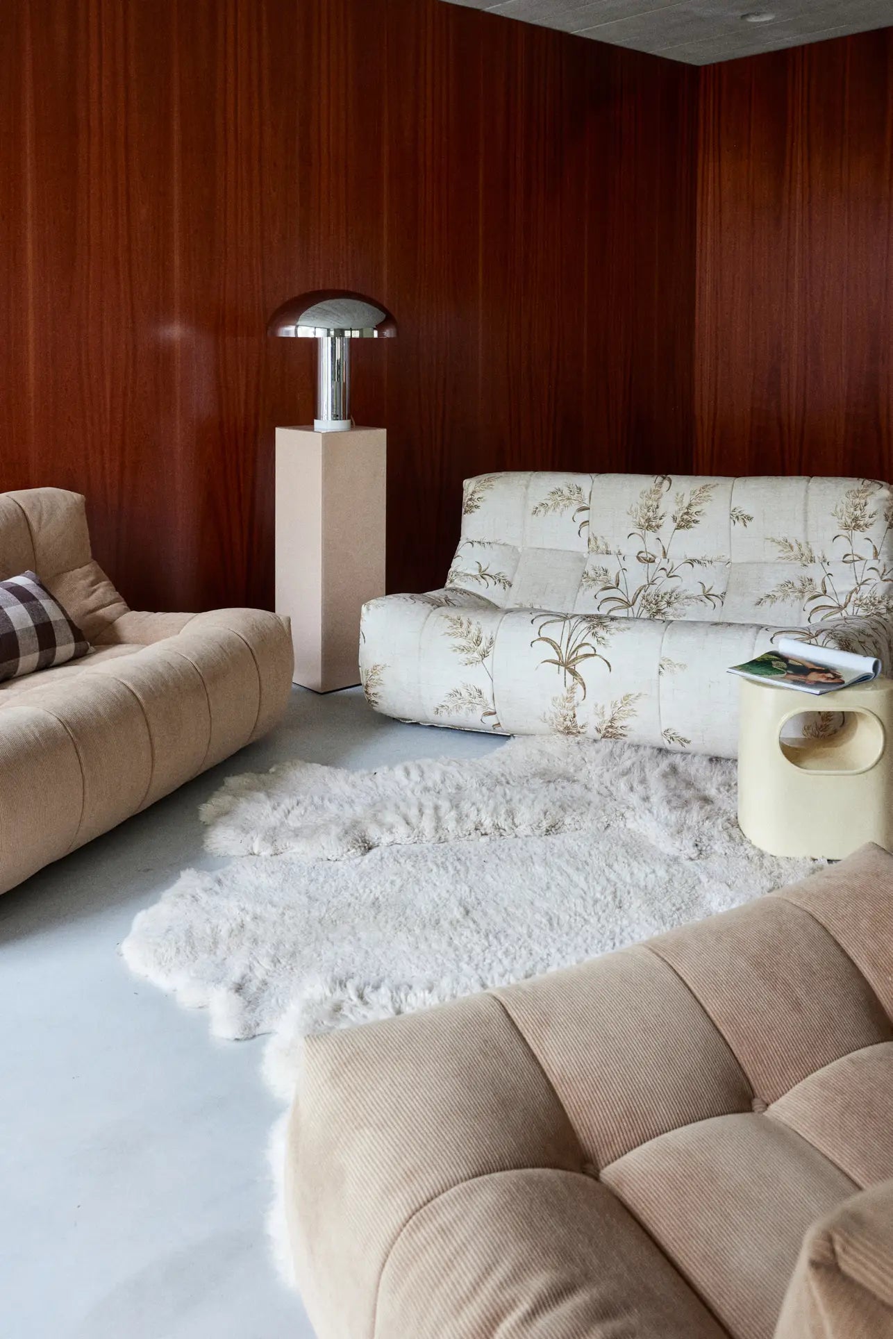 A cozy living room features tan corduroy sofas, a white floral sofa, a fluffy rug, books on a cream side table, and the HKLIVING Lounge Bench Corduroy by HKLIVING beside a modern lamp and wood-paneled walls.