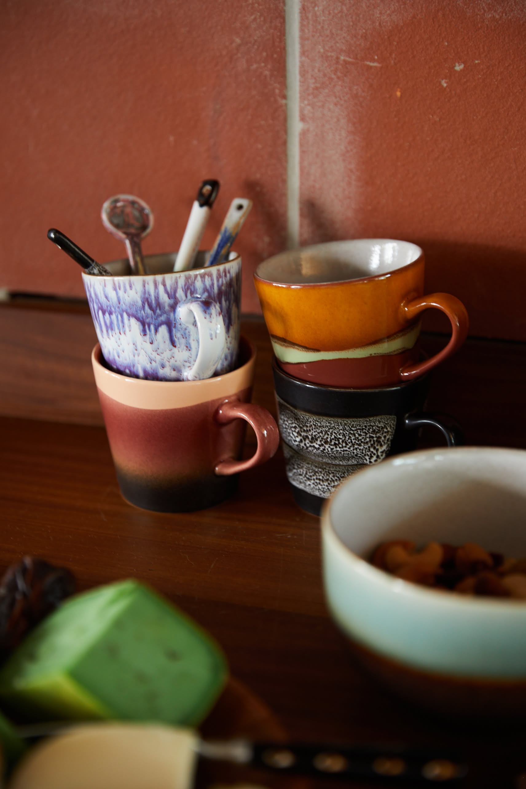 A stack of HKLIVING 70s Ceramics: Americano Mugs, Set of 4 sits on a wooden surface with several spoons, a bowl of mixed nuts, and a green object partially visible in front of a tiled wall.