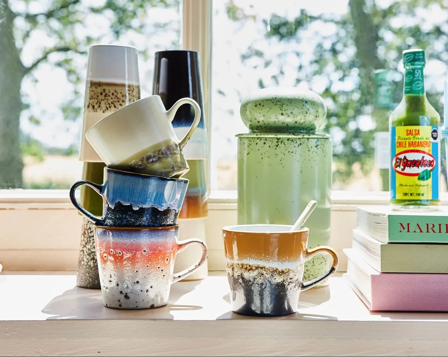 A set of four HKLIVING 70s Ceramics: Americano Mugs sits stacked on a windowsill among vases, a green jar, hot sauce, and books, sunlight highlighting their colorful, speckled design and retro style.