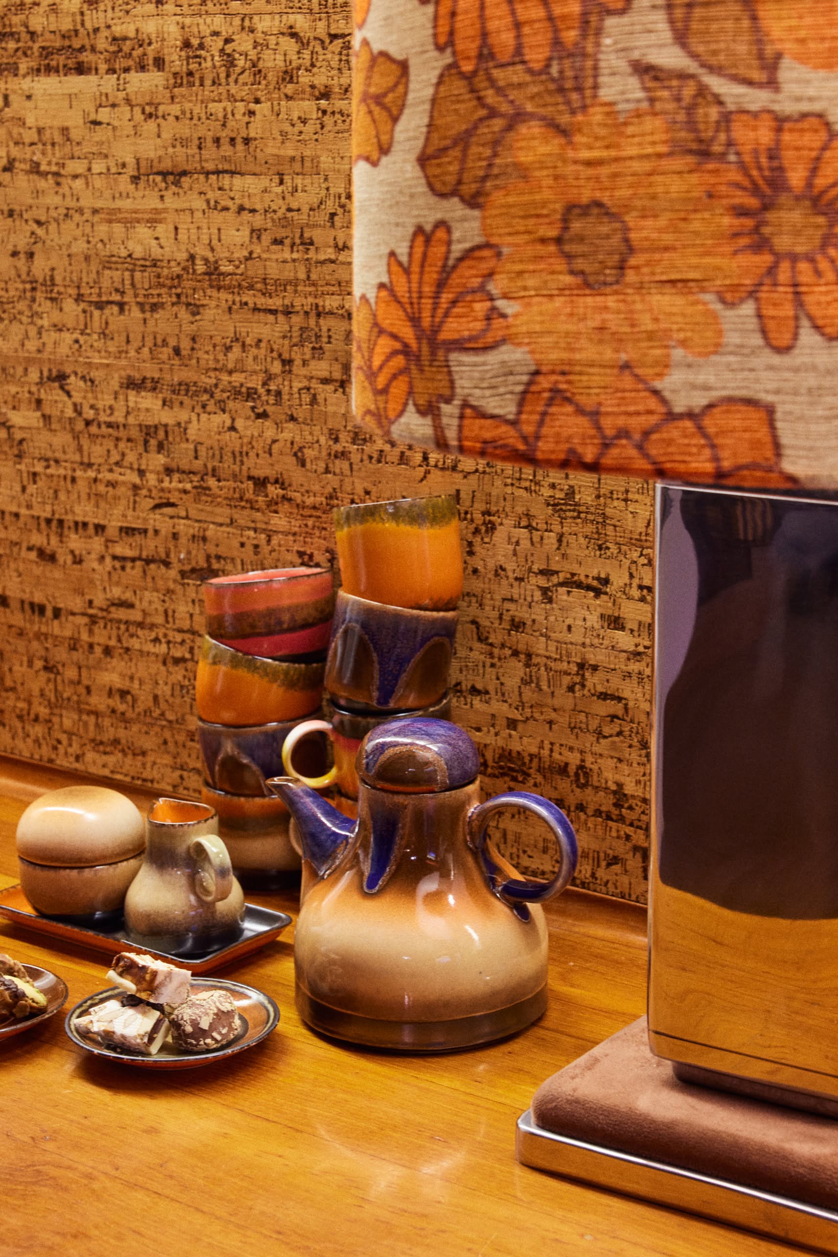 A retro scene with HKliving 70s Ceramics: Café Cups (Set of 4 - Java) sits stacked on a wooden table. An orange floral lampshade and cork wall complete the vintage 1970s vibe.