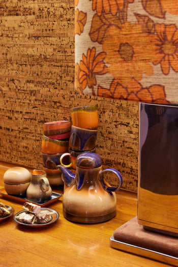 A retro scene with HKliving 70s Ceramics: Café Cups (Set of 4 - Java) sits stacked on a wooden table. An orange floral lampshade and cork wall complete the vintage 1970s vibe.