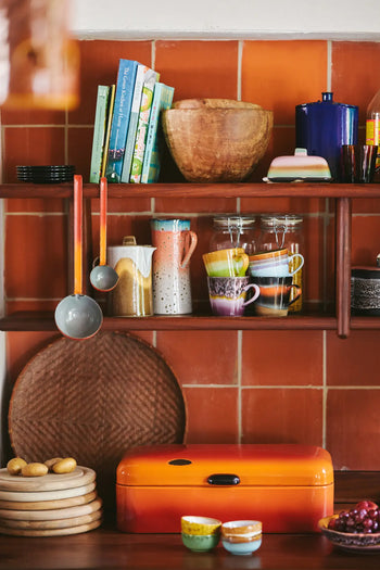 A kitchen shelf displays cookbooks, wooden bowls, HKLIVING 70s Ceramics: Cappuccino Mugs (Set of 4), glass jars, and ladles. Below sit a round orange breadbox, stacked plates, potatoes, and small dishes against red-brown tiled walls.