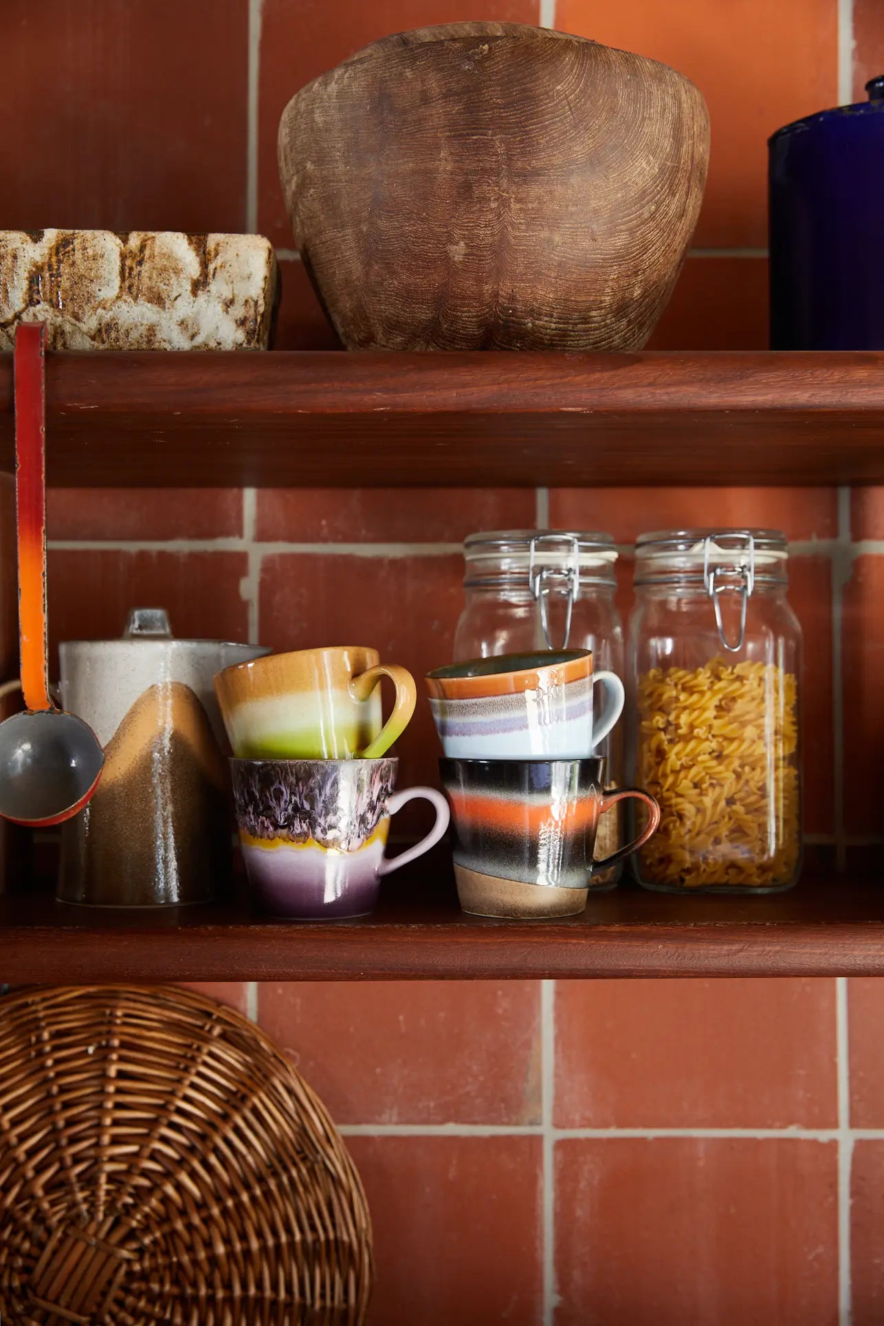 A kitchen shelf displays two HKLIVING 70s Ceramics Cappuccino Mugs, a sugar jar, a pasta jar, a metal scoop with red handle, a wicker tray, rustic bowl, and another container—all set against a tiled wall.