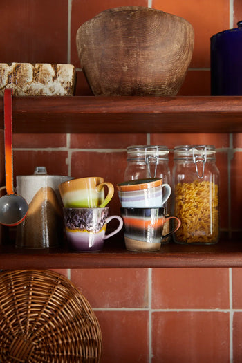 A kitchen shelf displays two HKLIVING 70s Ceramics Cappuccino Mugs, a sugar jar, a pasta jar, a metal scoop with red handle, a wicker tray, rustic bowl, and another container—all set against a tiled wall.