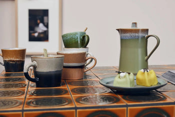 On a patterned tile table, the HKLIVING 70s Ceramics: Cappuccino Mugs, Set of 4 rest in earthy tones near a ceramic coffee pot, saucers, and a plate of green and yellow desserts; their hand-glazed finish channels vintage ‘70s style.