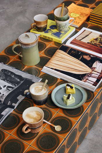 A retro-style table with a geometric orange pattern holds HKLIVING 70s Ceramics: Cappuccino Mugs (Set of 4), a green teapot, books, magazines, and a plate with two pastries topped with chocolate pieces.