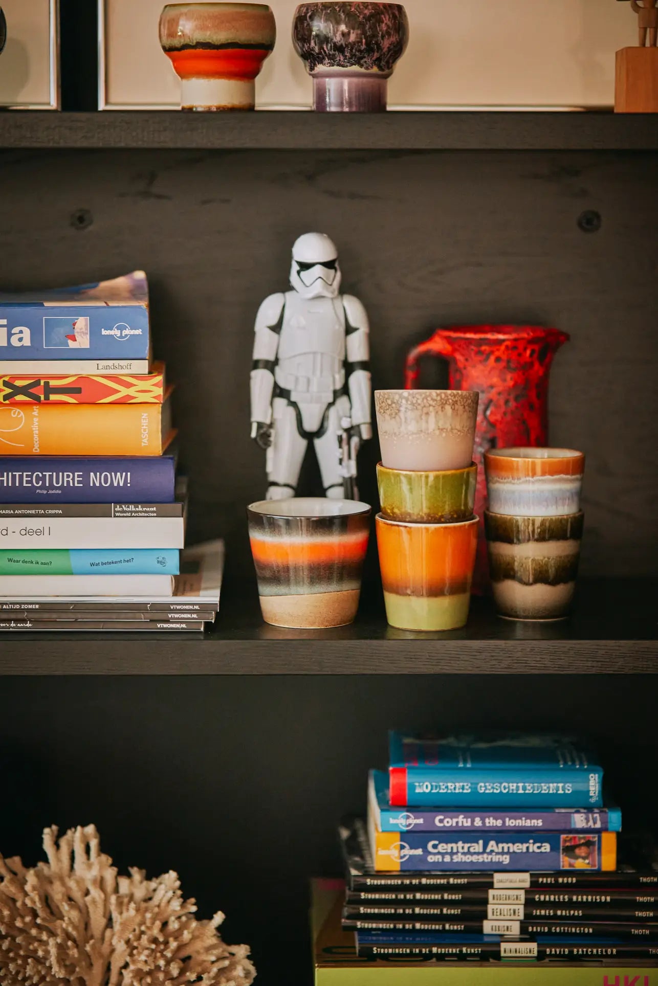 A black shelf displays HKLIVING 70s Ceramics Coffee Mugs (Set of 6), a red pitcher, white Stormtrooper figurine, and books on architecture, travel, and design. A piece of coral rests on the lower shelf.
