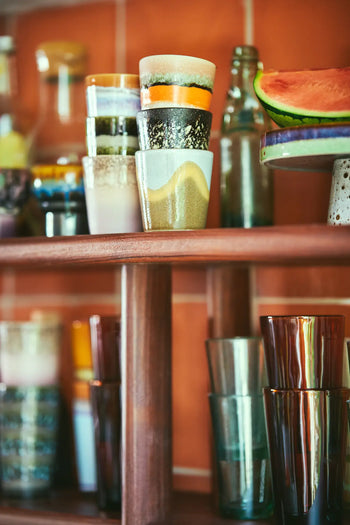 A wooden kitchen shelf displays the HKLIVING 70s Ceramics Coffee Mugs (Set of 6) alongside assorted glassware, bottles, and a plate with watermelon, all set against vibrant orange tiles.