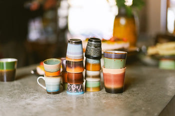 The HKLIVING 70s Ceramics: Coffee Mugs, Set of 6, features colorful handcrafted glazing and sits on a gray countertop in a softly lit kitchen, with blurred dishes and vintage decor in the background.