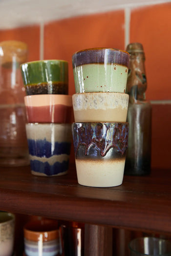 A set of six HKLIVING 70s Ceramics coffee mugs, handcrafted with earth-toned glazes, is stacked on a wooden shelf against a reddish tile wall, capturing vintage charm.