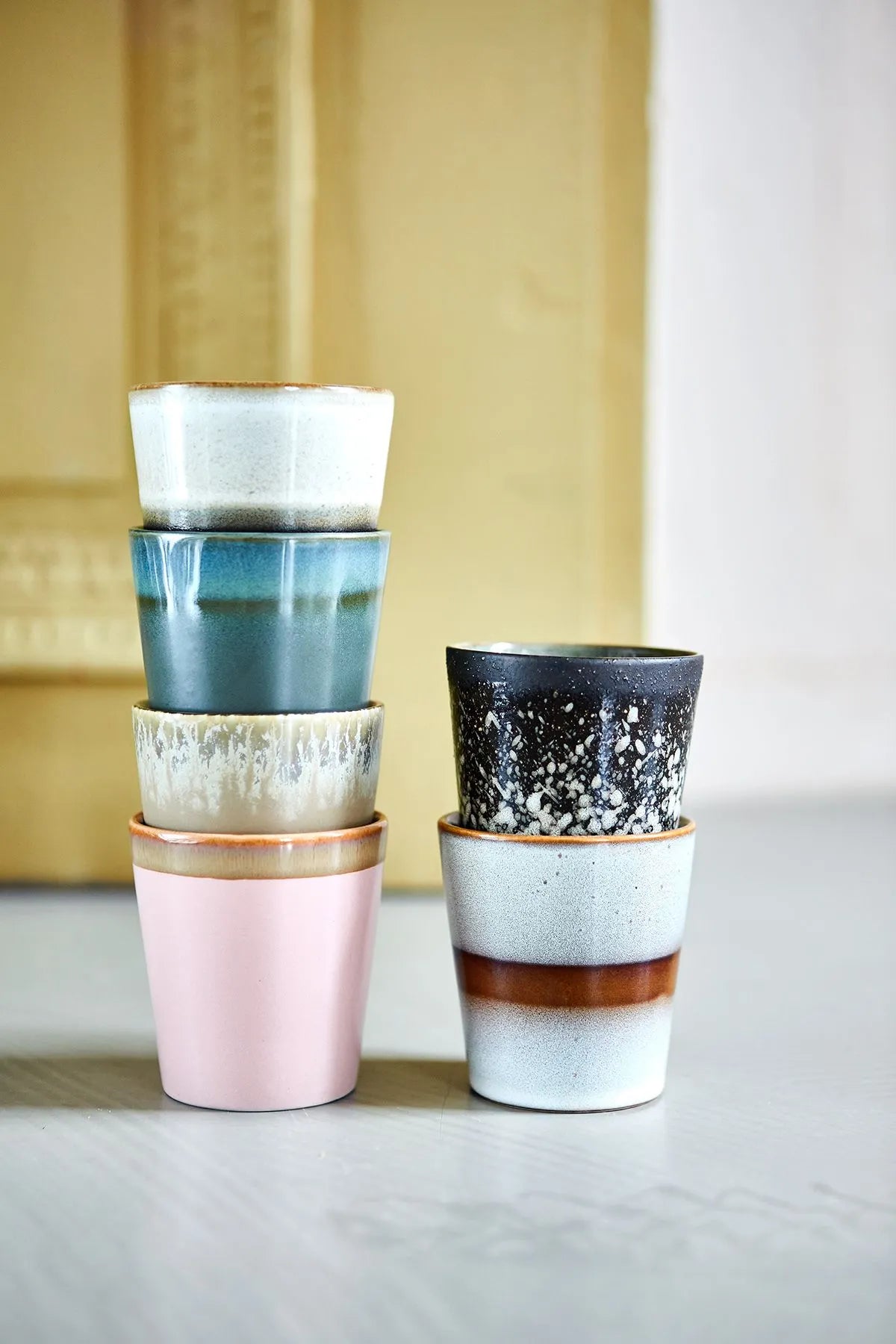 The HKLIVING 70s Ceramics Coffee Mugs, Set of 6, features handcrafted glazing and unique designs—pastel pink, blue, black with white speckles, and brown stripes—stacked in two uneven columns to showcase vintage-inspired charm.