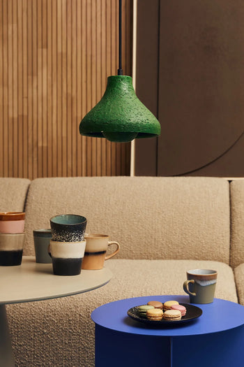 A cozy seating area with a beige textured sofa, a table displaying HKLIVING 70s Ceramics: Coffee Mugs (Set of 6), a blue side table holding a cup and macarons, and a green pendant light hanging above.
