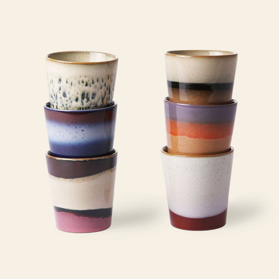 Six HKLIVING 70s Ceramics Coffee Mugs are stacked in two columns of three, each featuring a unique, colorful abstract design in stripes and gradients with blue, purple, cream, brown, and red on a light background.