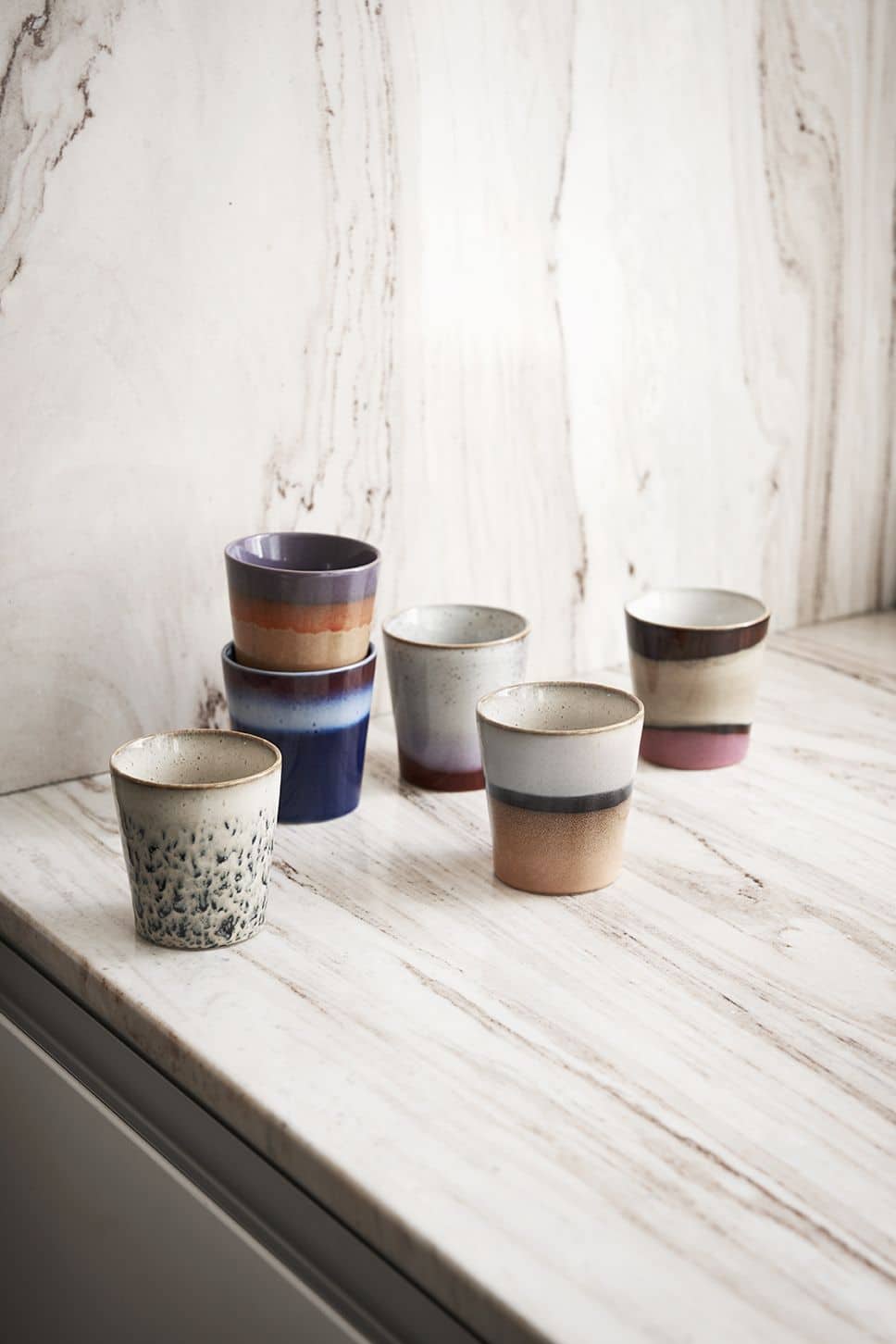Six HKLIVING 70s Ceramics Coffee Mugs, each with unique handcrafted glazing, are stacked in pairs on a marble countertop and backsplash, bringing retro charm to the bright, modern space.